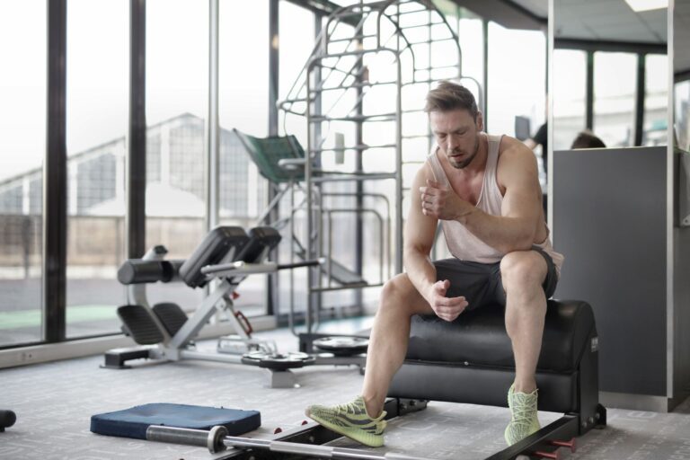 Tired young muscular male athlete in casual sportswear and sneakers sitting on bench and resting after intense weightlifting workout in modern fitness club