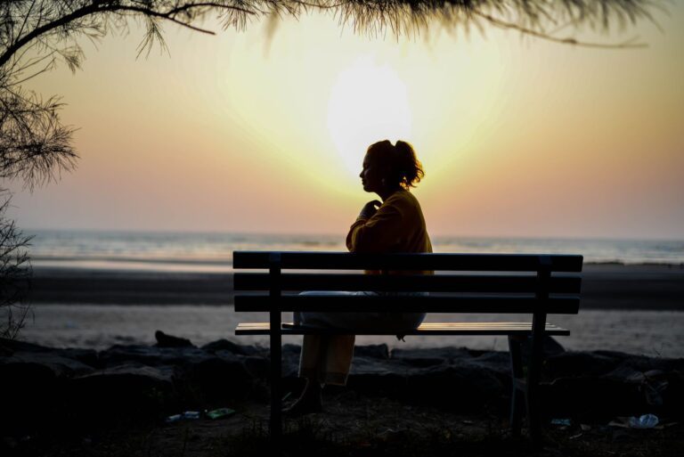 A silhouette of a woman sitting on a bench by the beach during sunrise, capturing solitude and contemplation.
