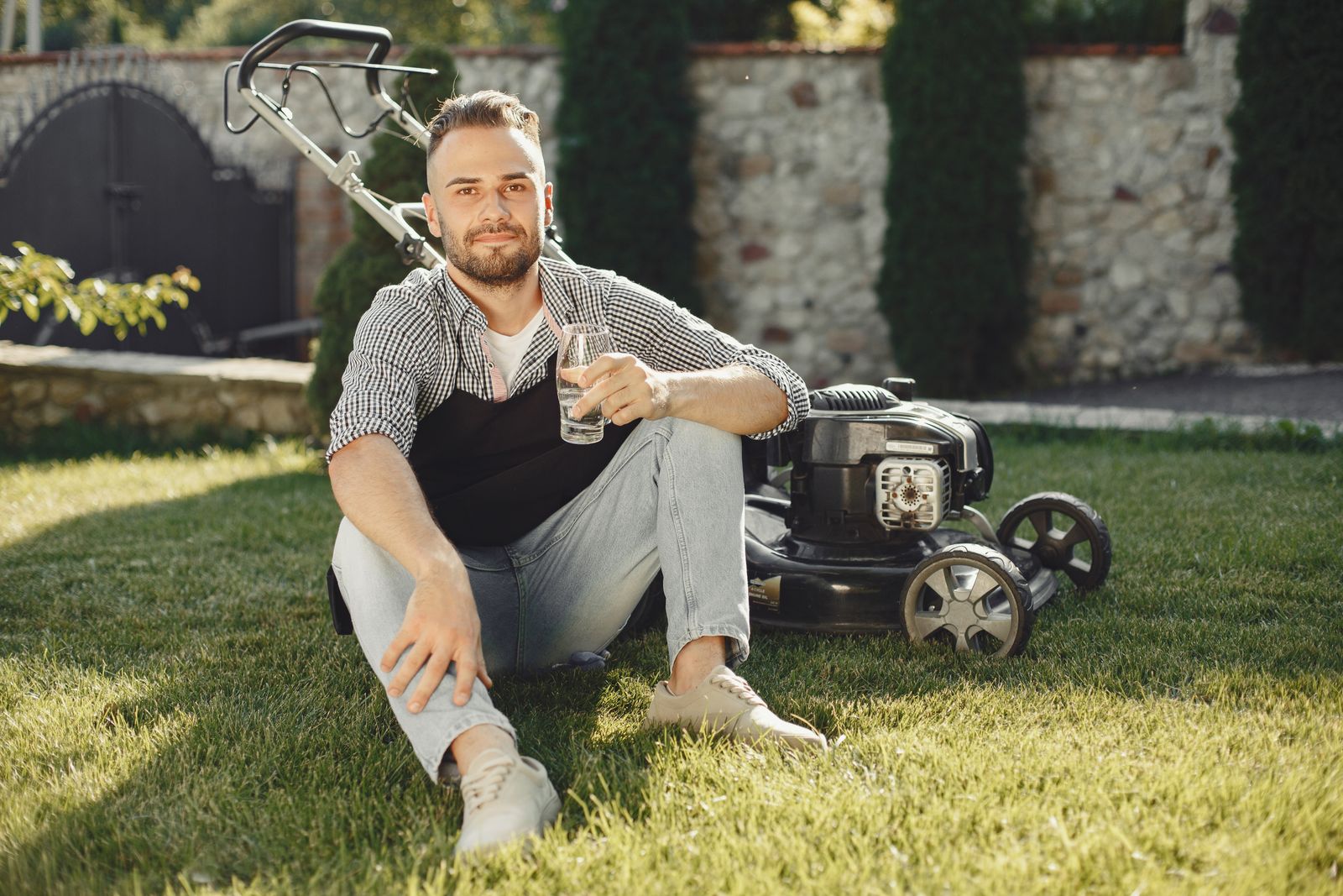 Man sitting on grass with drink, beside lawn mower on a sunny day.