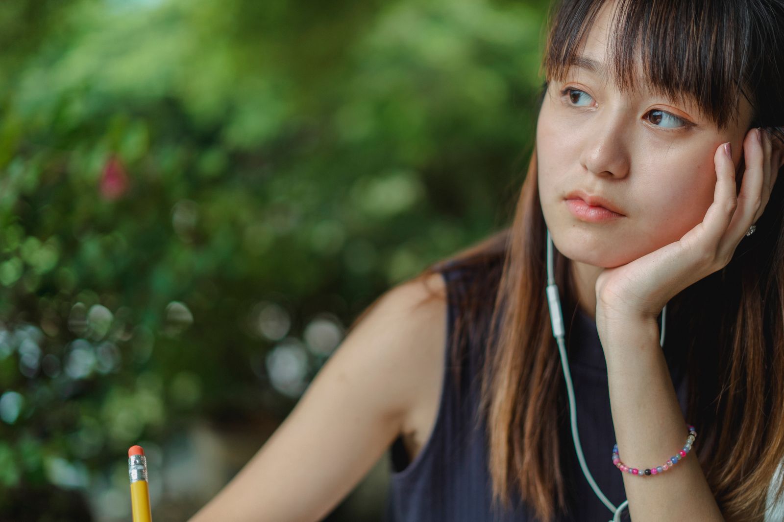 Pensive Asian female student listening to music via earphones while doing homework and taking notes with pencil