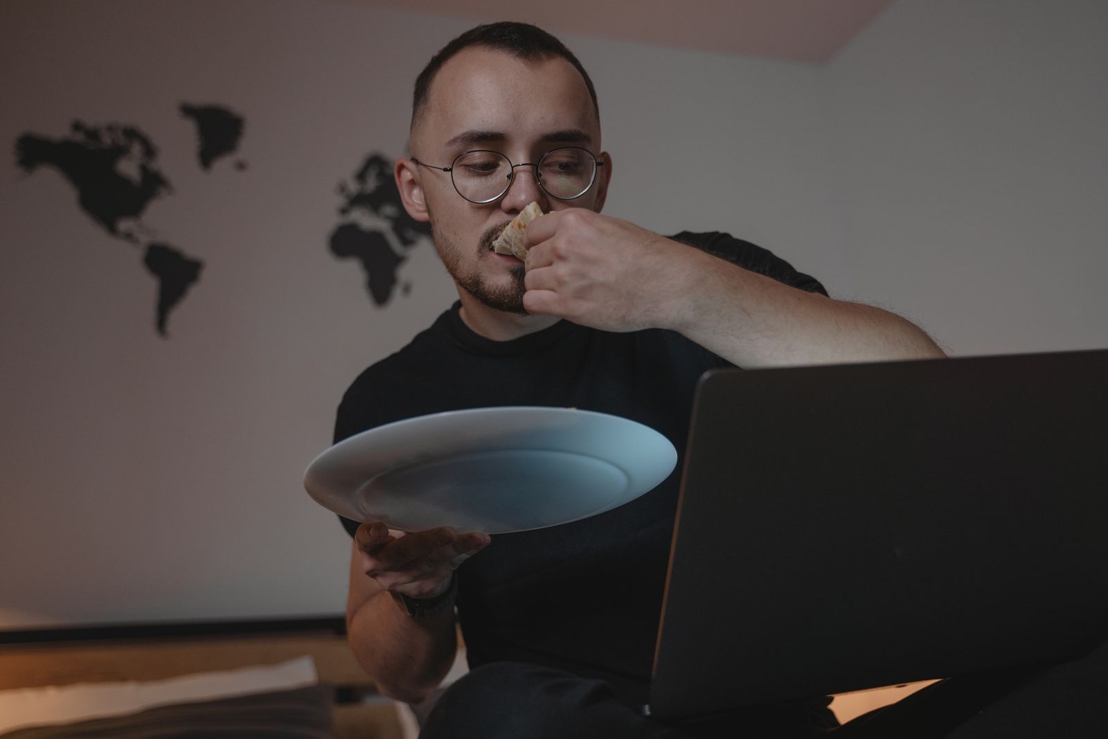 Man eating dinner while working on a laptop in an apartment with a world map on the wall.