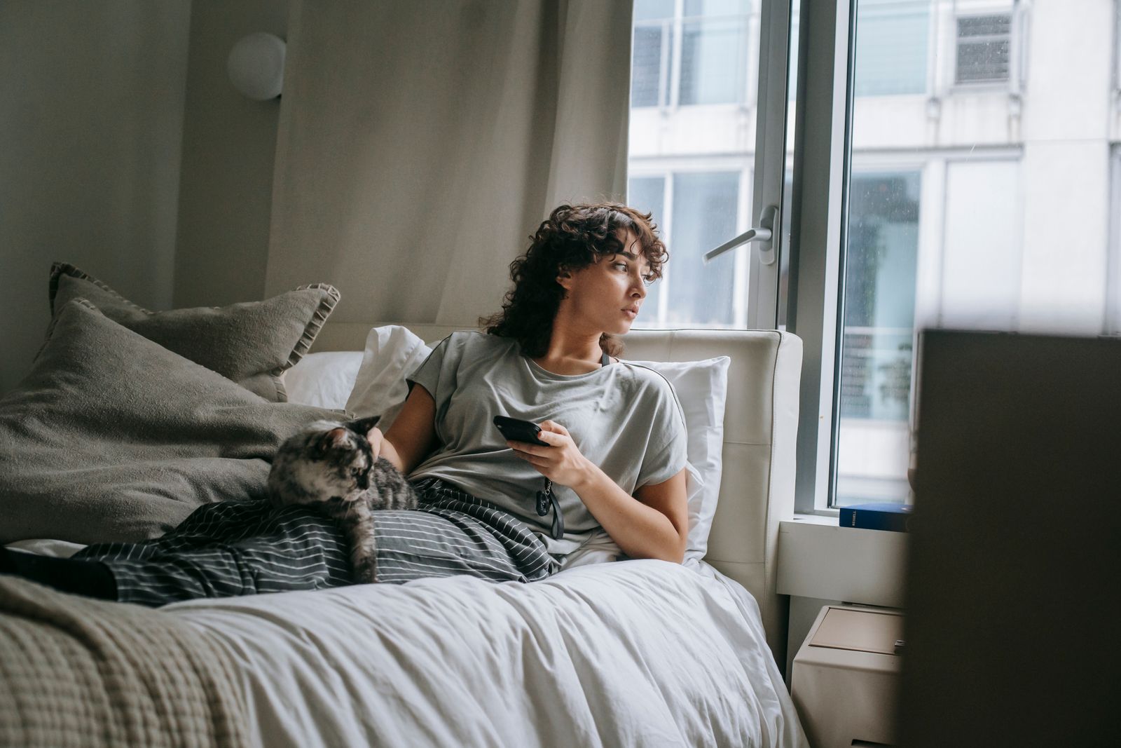 Full body of female owner looking at window and stroking cat while lying on bed and surfing smartphone on weekend