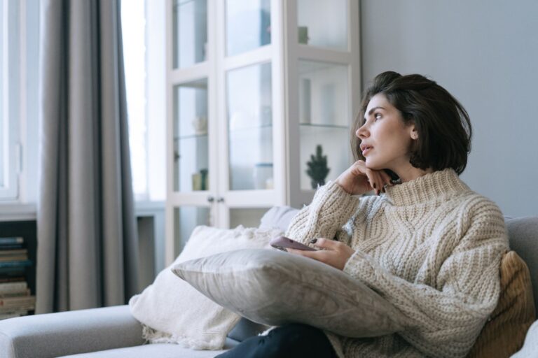 Woman in a comfy sweater sitting on couch, looking thoughtful with a smartphone in hand.