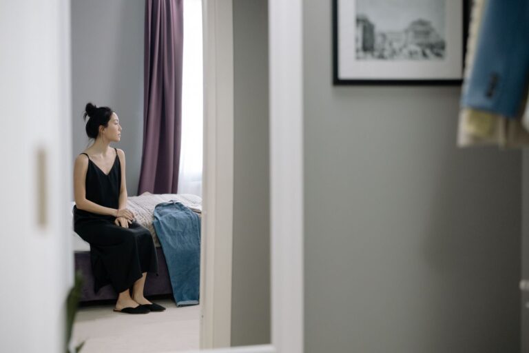 Woman in black dress sitting on bed in a minimalist bedroom setting, reflecting a calm and serene atmosphere.