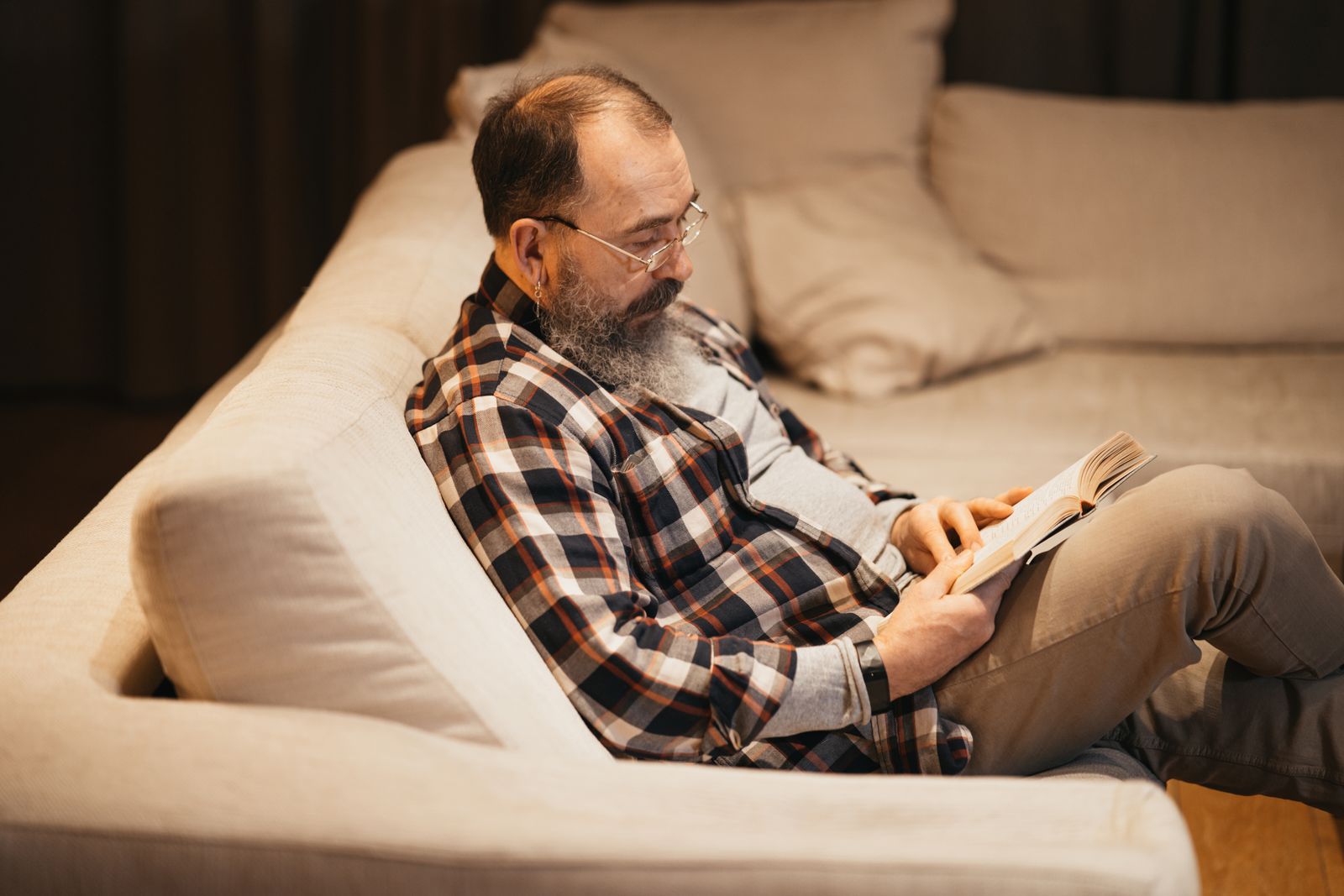 Adult bearded man wearing glasses, engrossed in a book, sitting on a cozy sofa in a warm indoor setting.