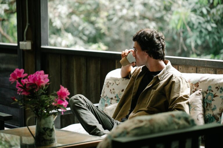 Adult male drinking aromatic hot beverage from glass while sitting on couch and looking away against blooming flowers on veranda