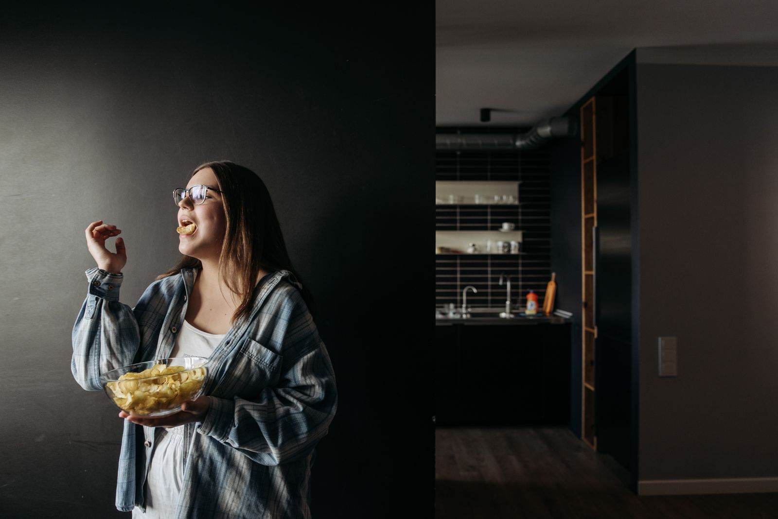 A woman in glasses enjoys chips indoors, standing in a modern kitchen environment.