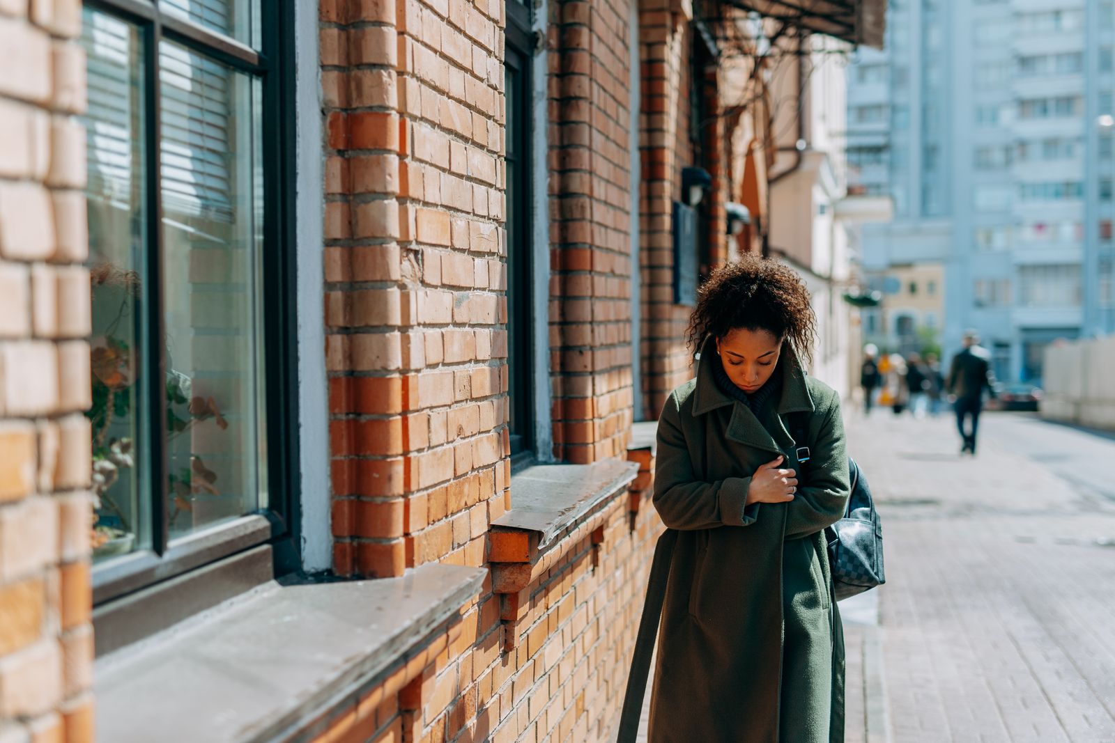 A woman in a green coat walking along a brick building on a sunny day, reflecting solitude.