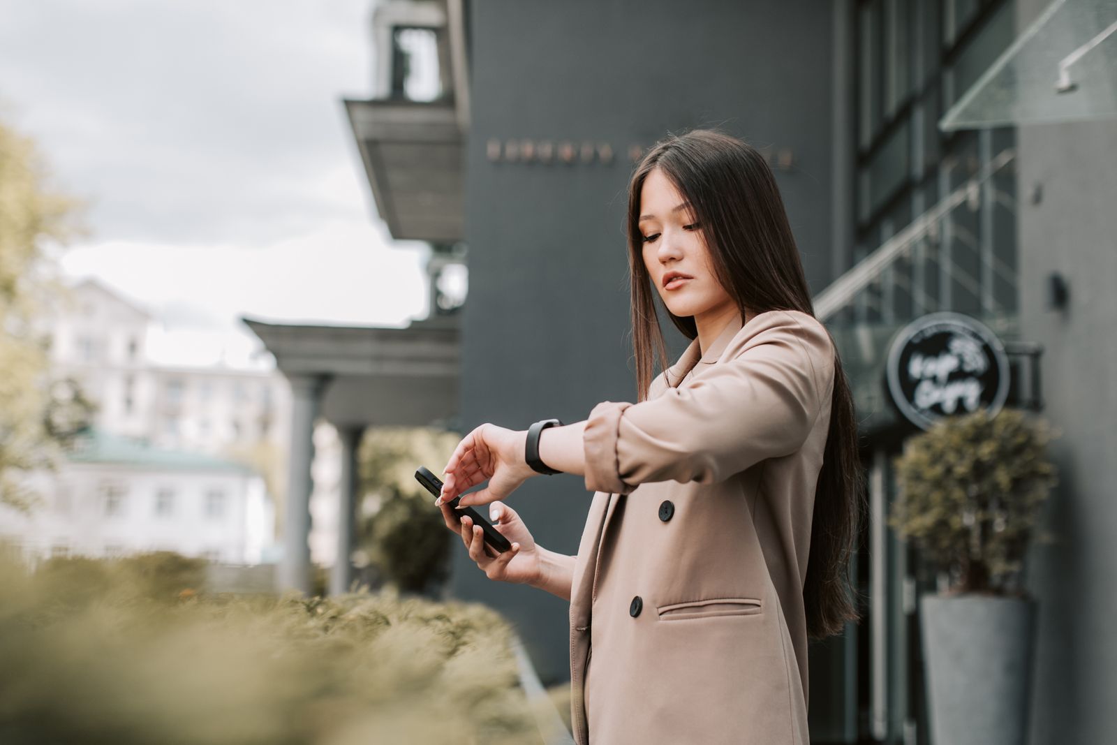 Businesswoman in corporate attire checking her smartwatch while holding a phone outdoors.