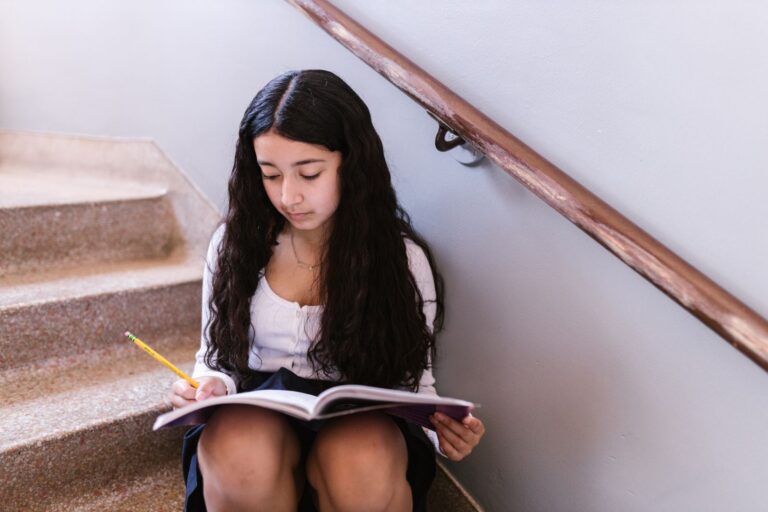 A focused teenage girl with long hair writing in a notebook on indoor stairs.