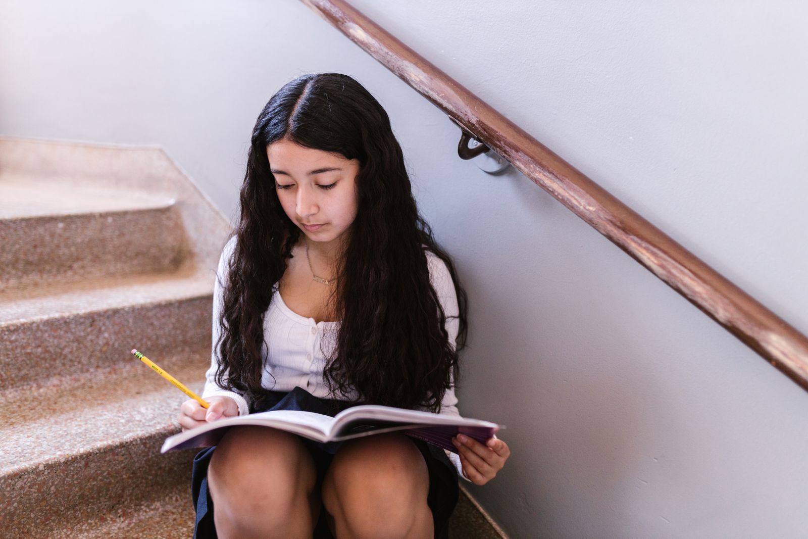 A focused teenage girl with long hair writing in a notebook on indoor stairs.