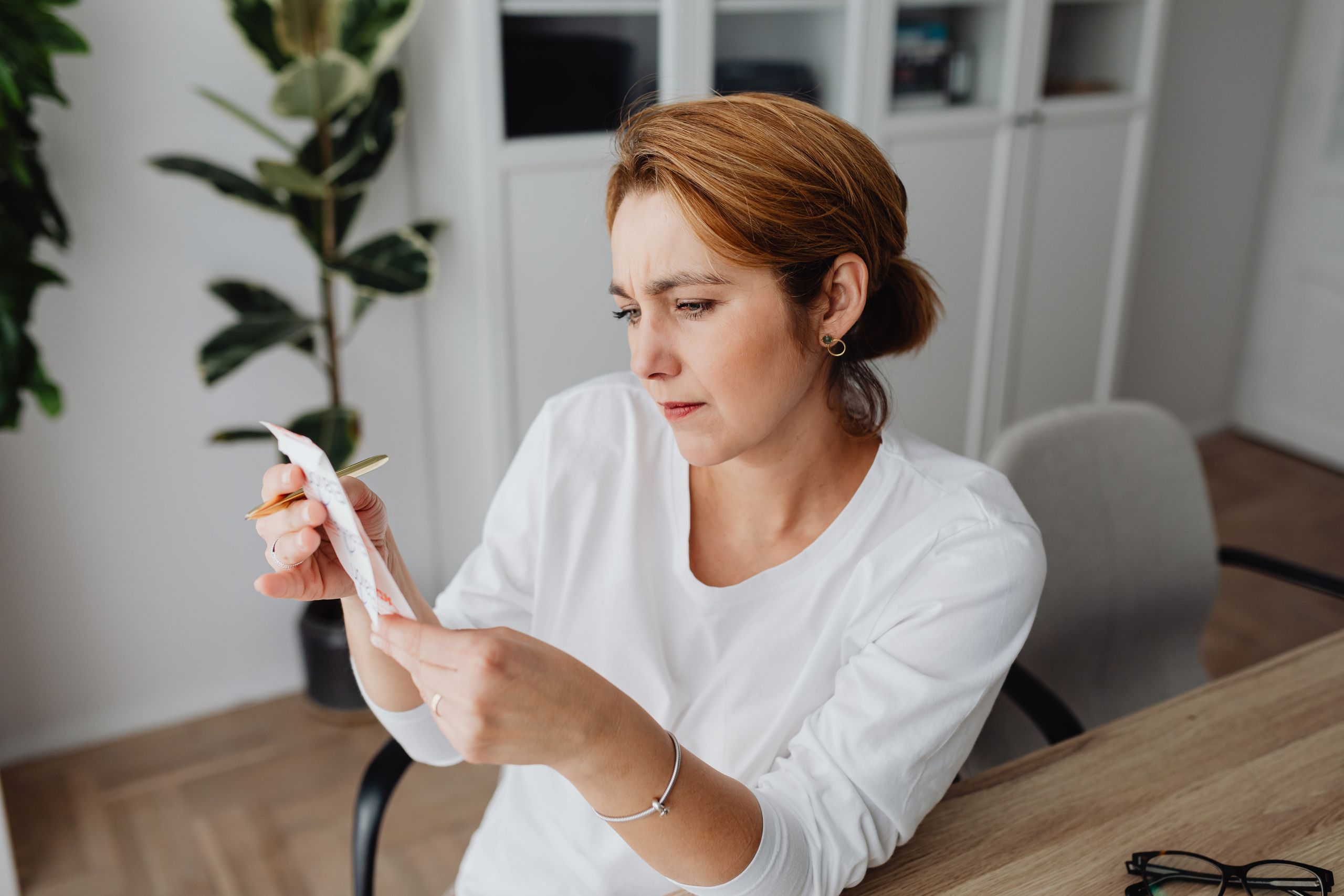 A concerned adult woman examines a receipt while sitting at her office desk, indicating financial scrutiny.