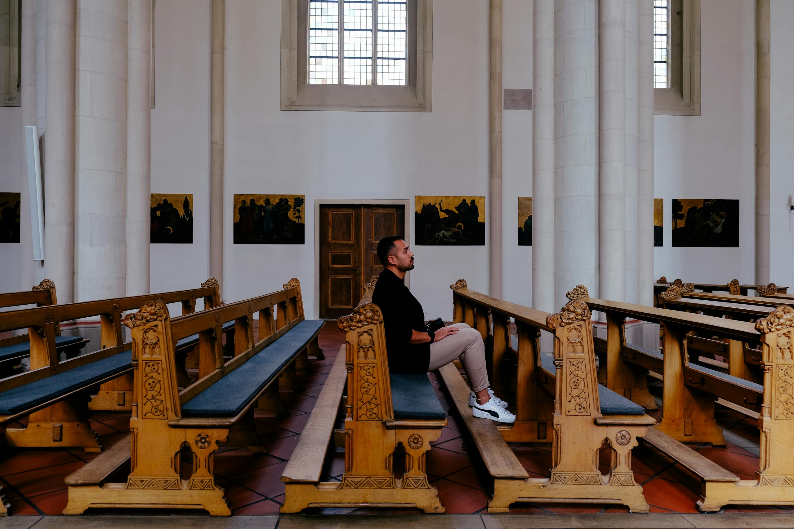 A man sits alone in a church pew, surrounded by ornate wooden benches and religious art.