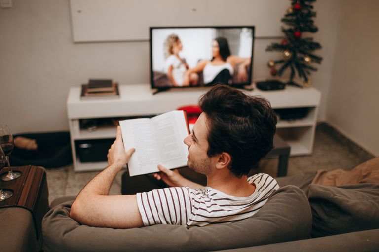 A man sits on a sofa reading a book in a cozy living room with a TV on.