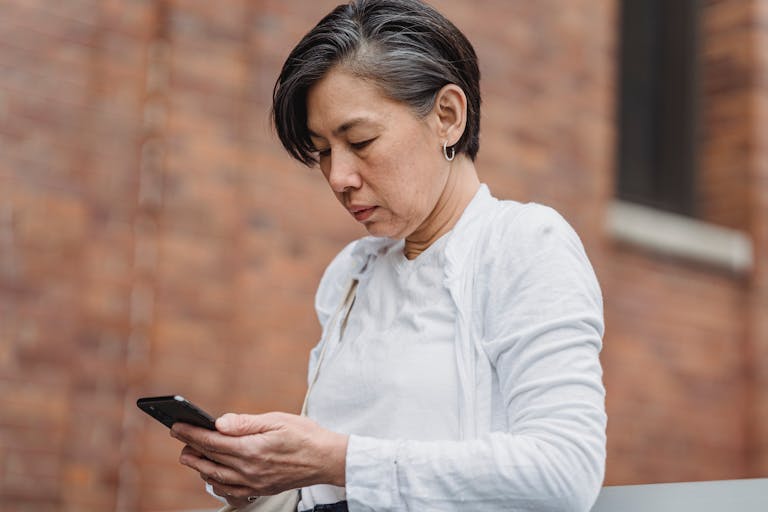 A middle-aged woman intently uses her smartphone against a brick wall backdrop.