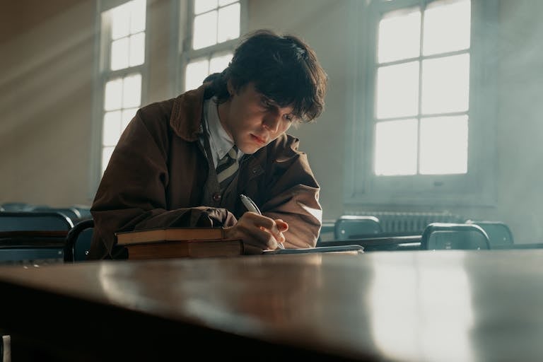 A student deeply engaged in writing at a desk, with books, in a sunlit classroom.
