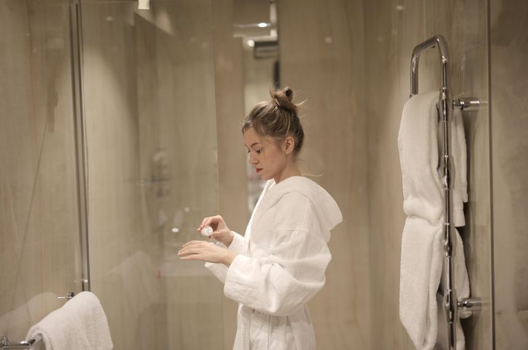 A woman in a bathrobe applies lotion in a contemporary bathroom with glass doors and towels.