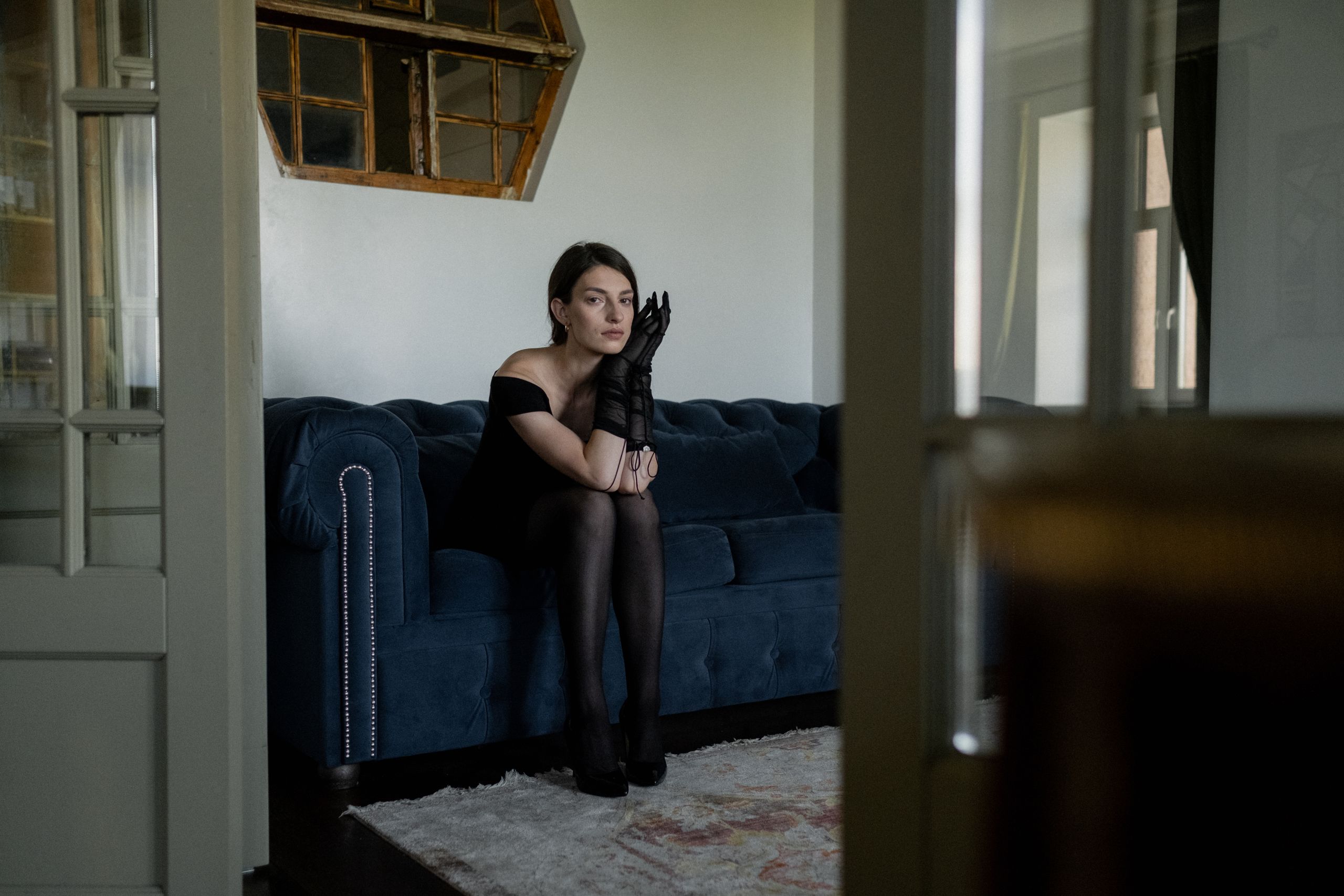 A woman in a black dress sits on a sofa, expressing introspection and sorrow.