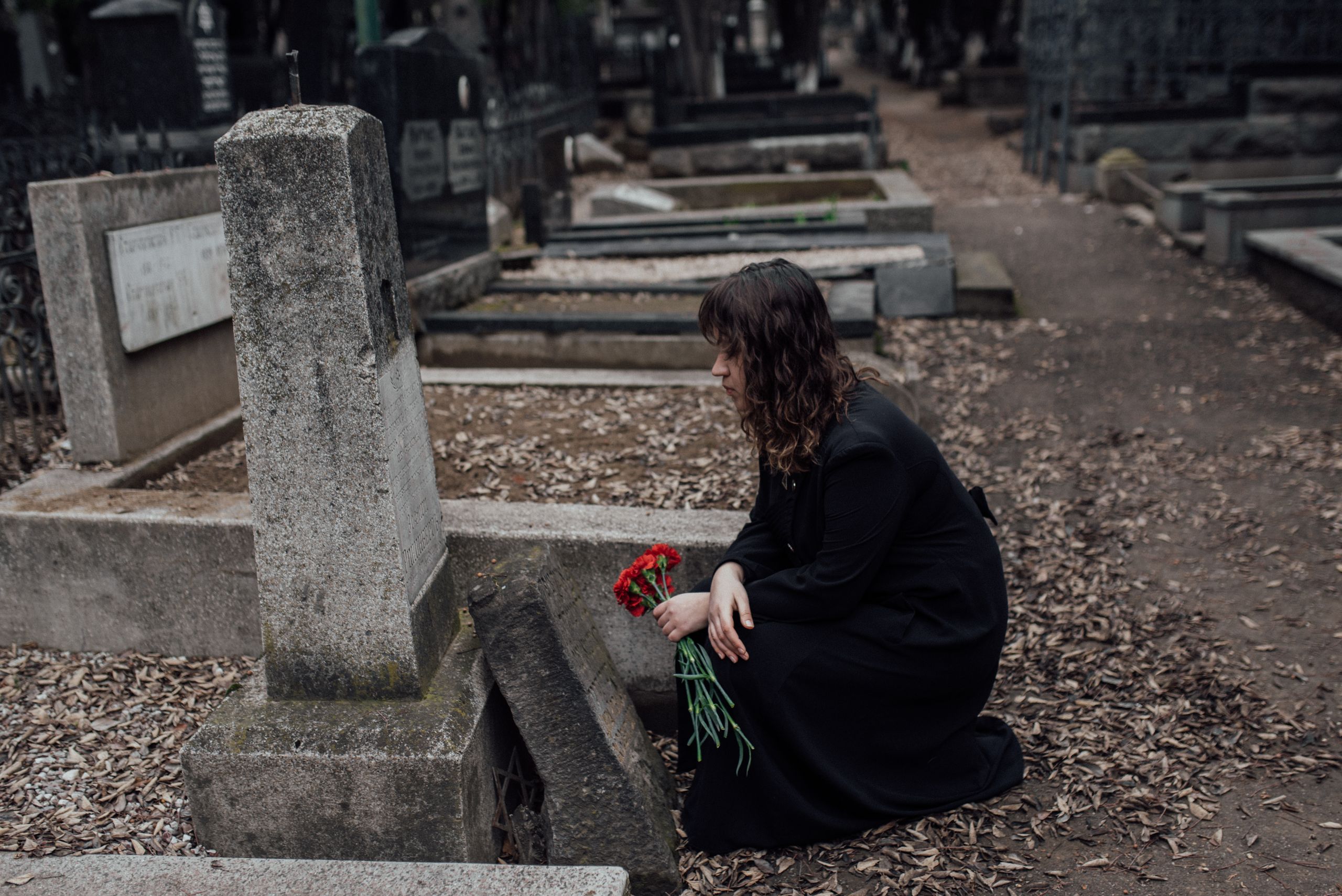 A woman in mourning holds red flowers in a quiet cemetery.