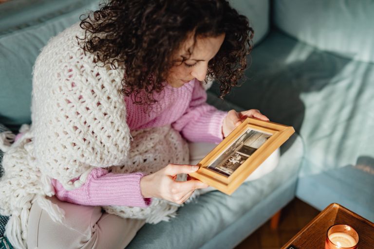 A woman with curly hair sitting indoors, gazing thoughtfully at a framed photo on the couch.