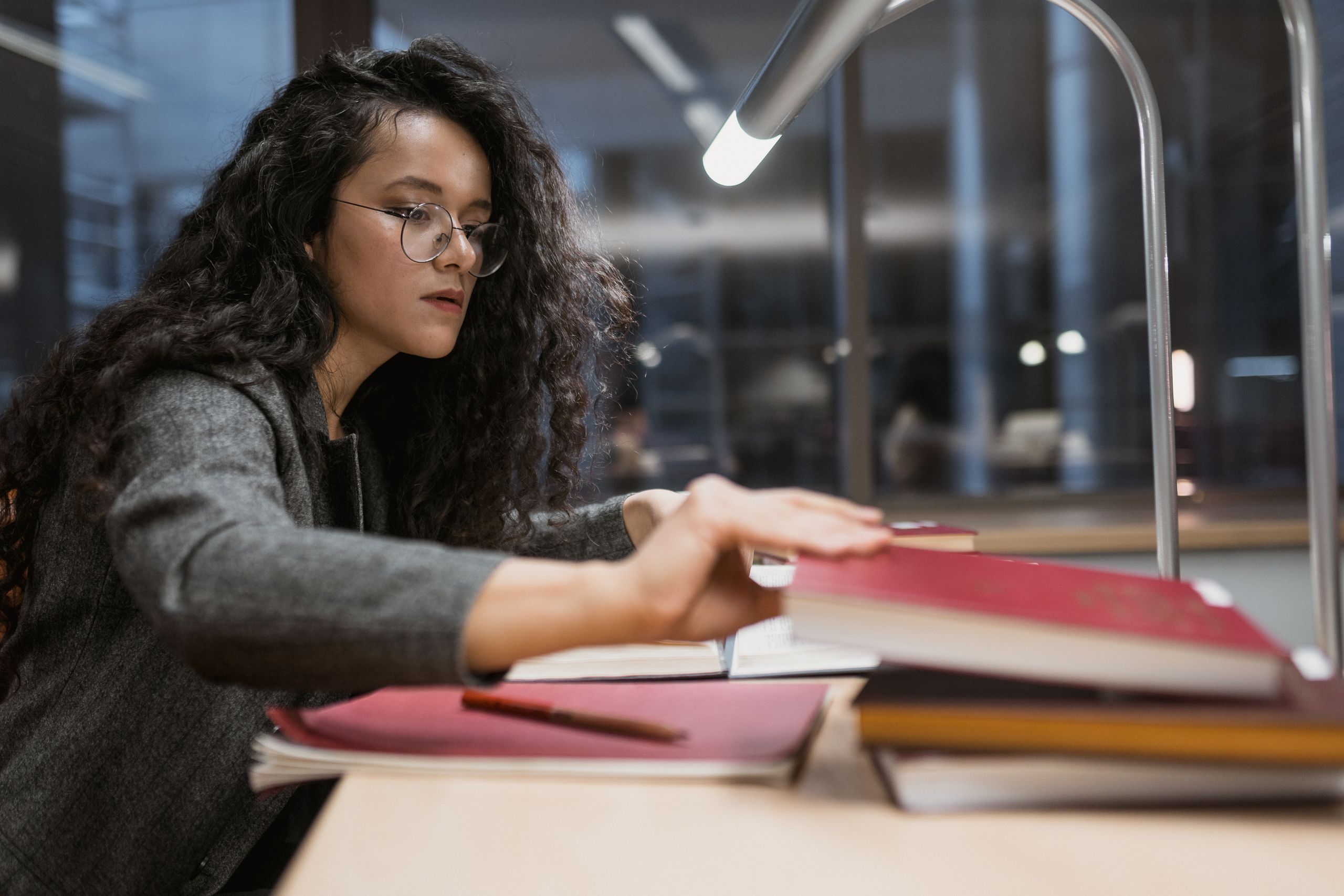 A woman with glasses studying at a desk under a lamp in a library at night.