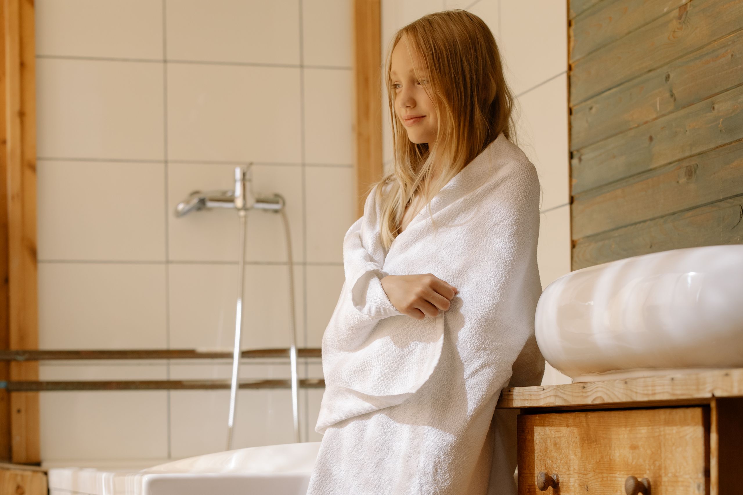 A young blonde girl wrapped in a towel standing by a bathroom sink, in a warm and cozy setting.
