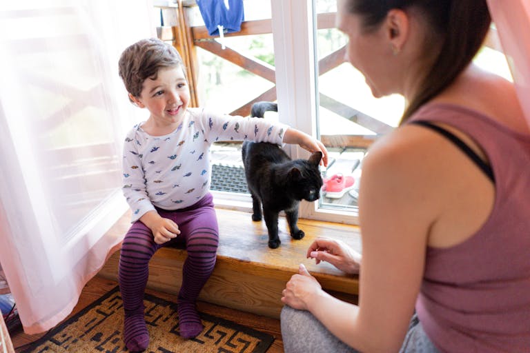 A young child smiling and playing with a black cat while a mother watches nearby.
