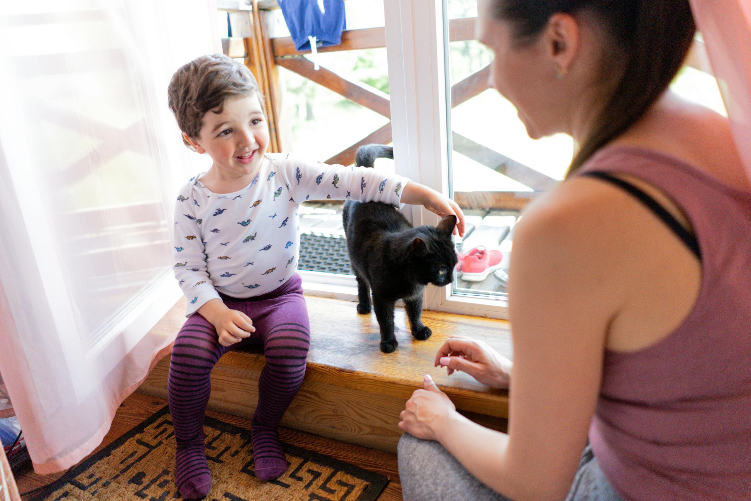 A young child smiling and playing with a black cat while a mother watches nearby.