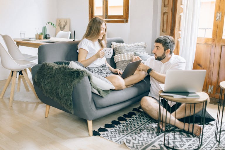 A young couple discussing work while using laptops in a cozy living room setting.