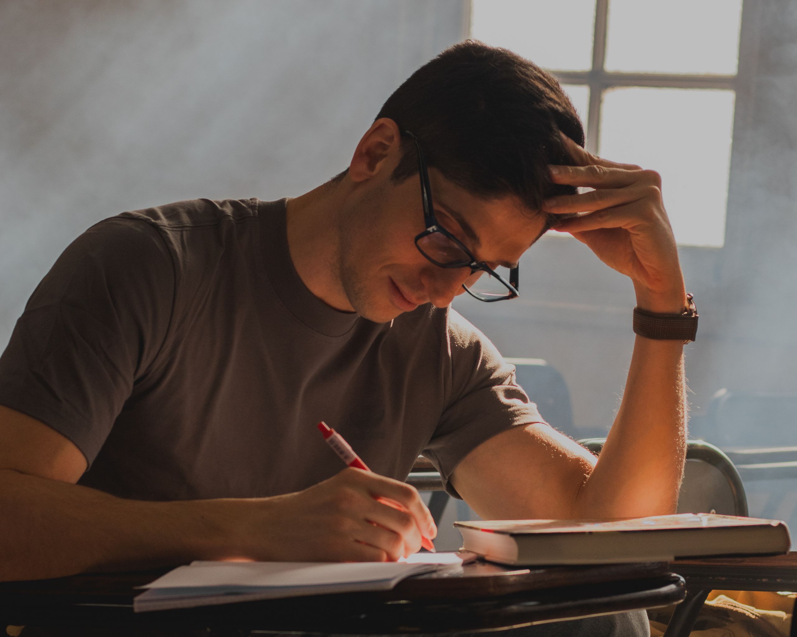 A young man with glasses intensely focused on writing in a notebook in a dim classroom.