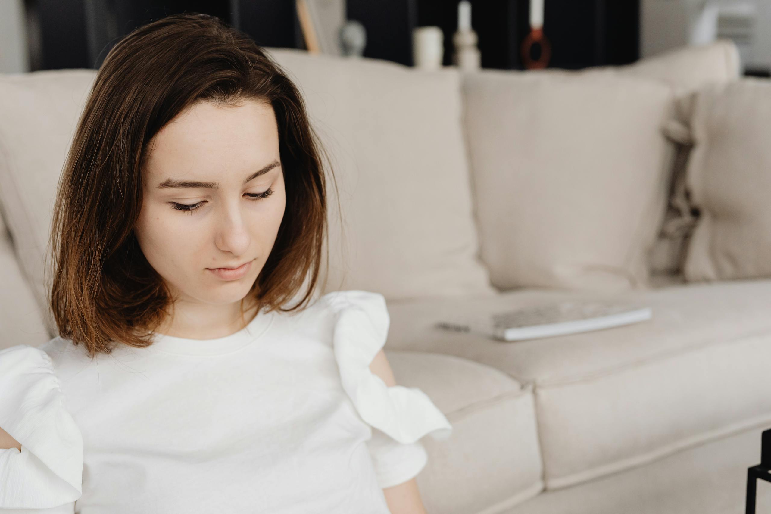 A young woman in a white top sits on a beige couch looking down, creating a serene indoor scene.