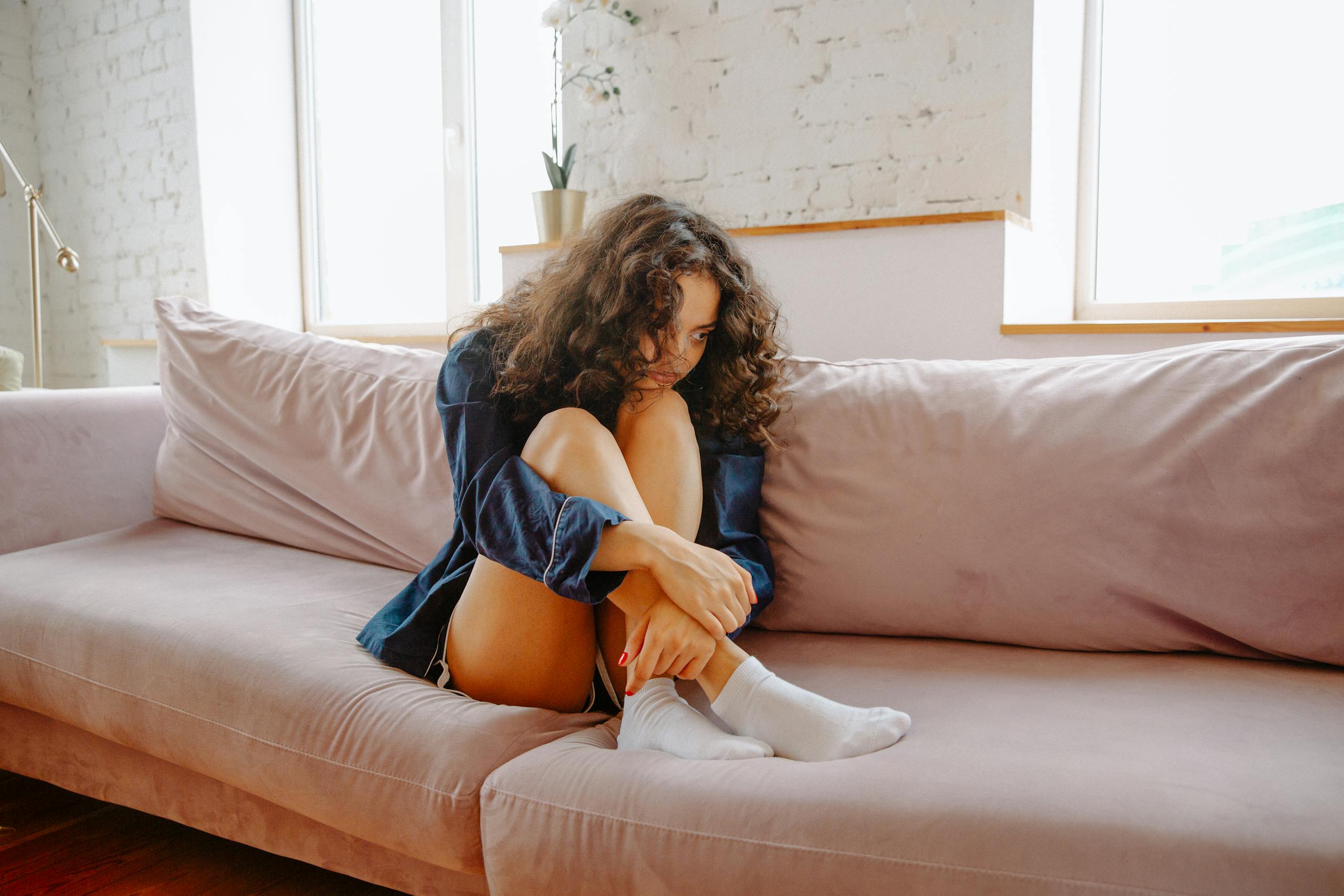 A young woman sits on a couch, appearing contemplative, in a cozy indoor setting.