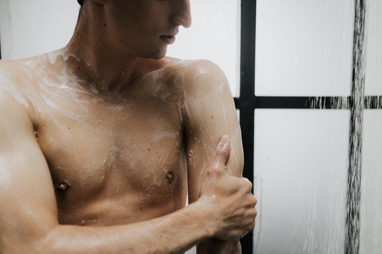 Adult man lathers soap on his body while showering, promoting cleanliness and fitness.