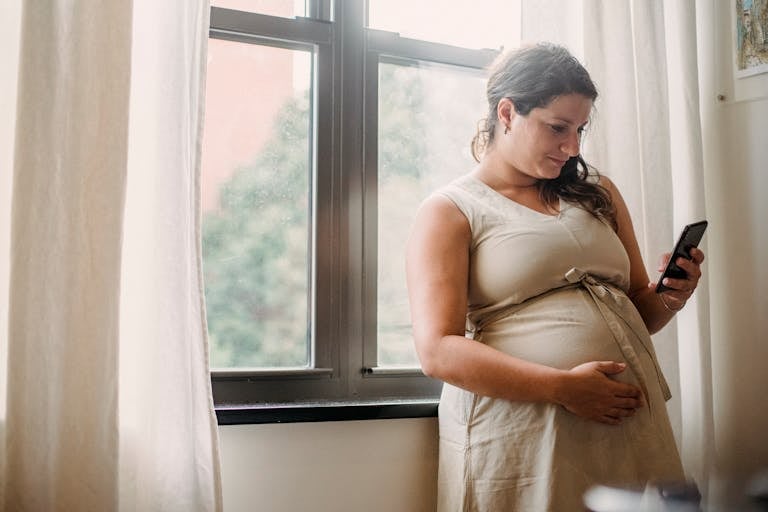 Adult pregnant lady checking information on phone while standing near window in room in daylight
