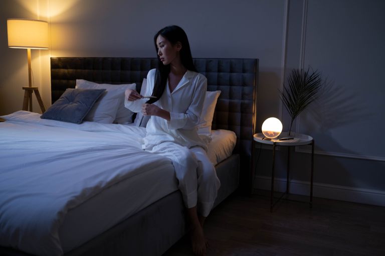 Asian woman in pajamas brushing hair on bed with warm lighting, creating a cozy atmosphere.