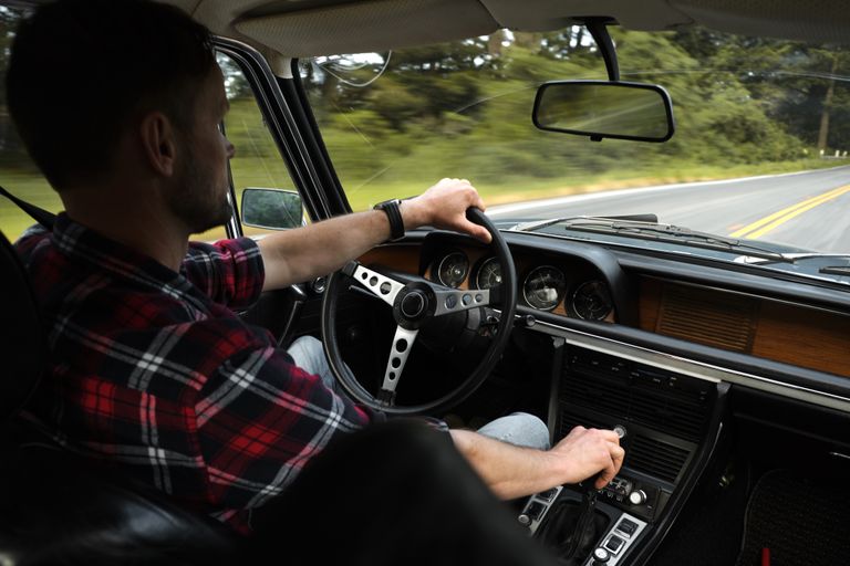 Back view of serious unshaven man in plaid shirt driving automobile by asphalt road among green trees