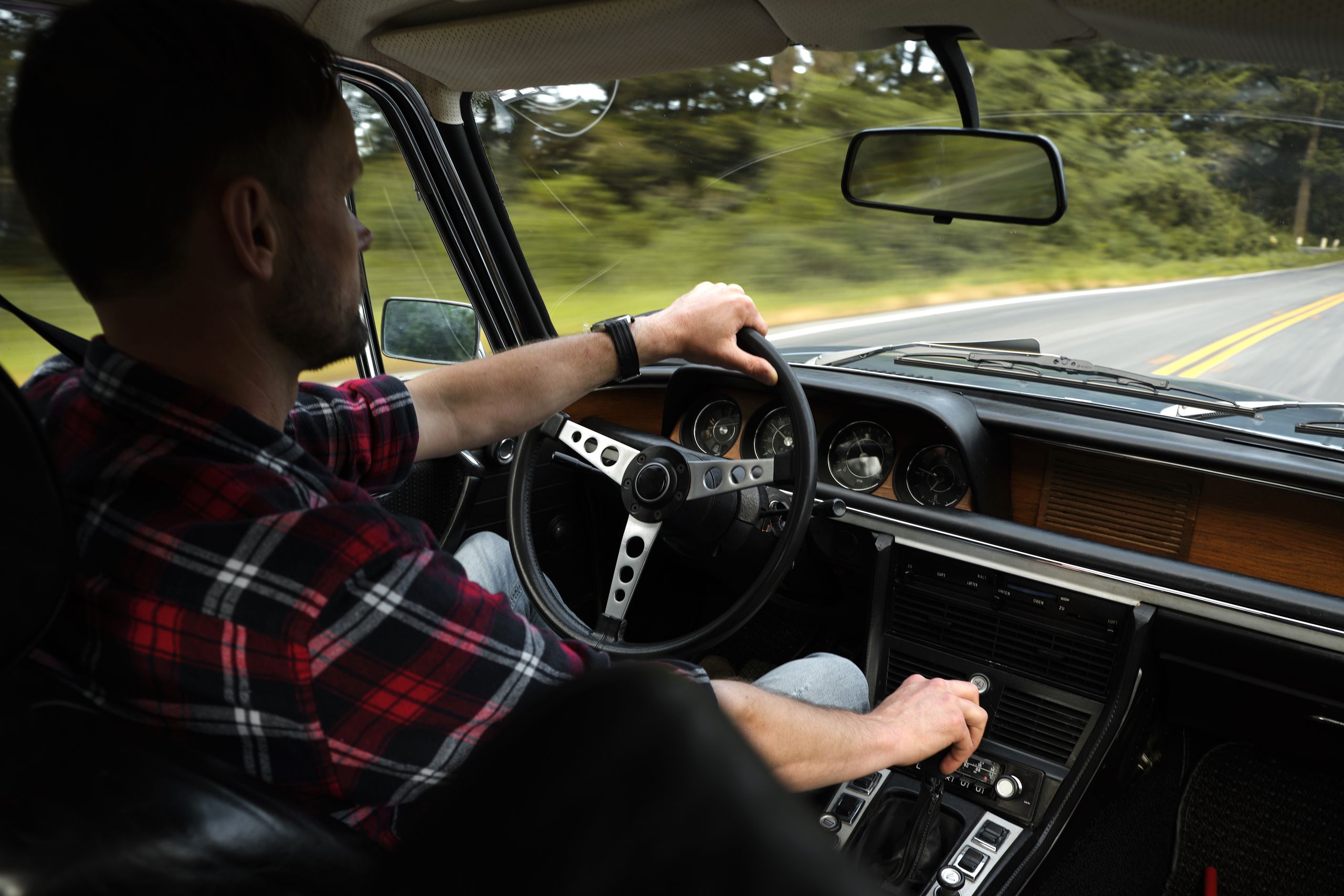 Back view of serious unshaven man in plaid shirt driving automobile by asphalt road among green trees