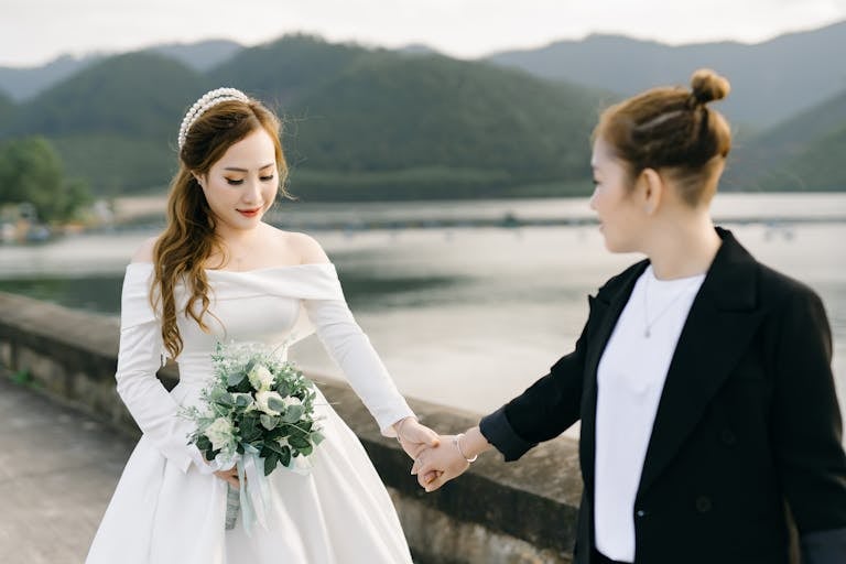 Bride in elegant gown holds bouquet while holding hands outdoors by a scenic lake.