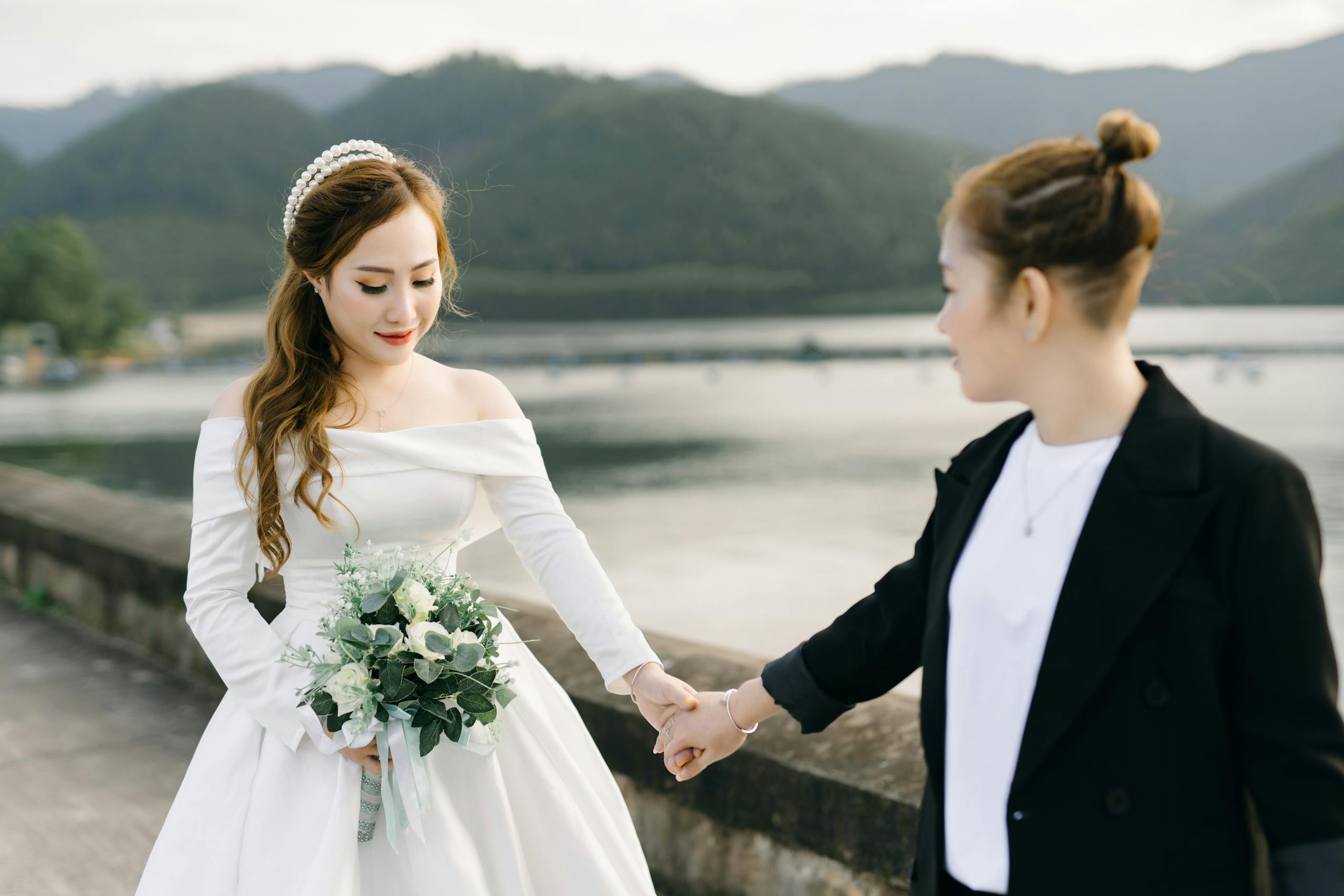 Bride in elegant gown holds bouquet while holding hands outdoors by a scenic lake.