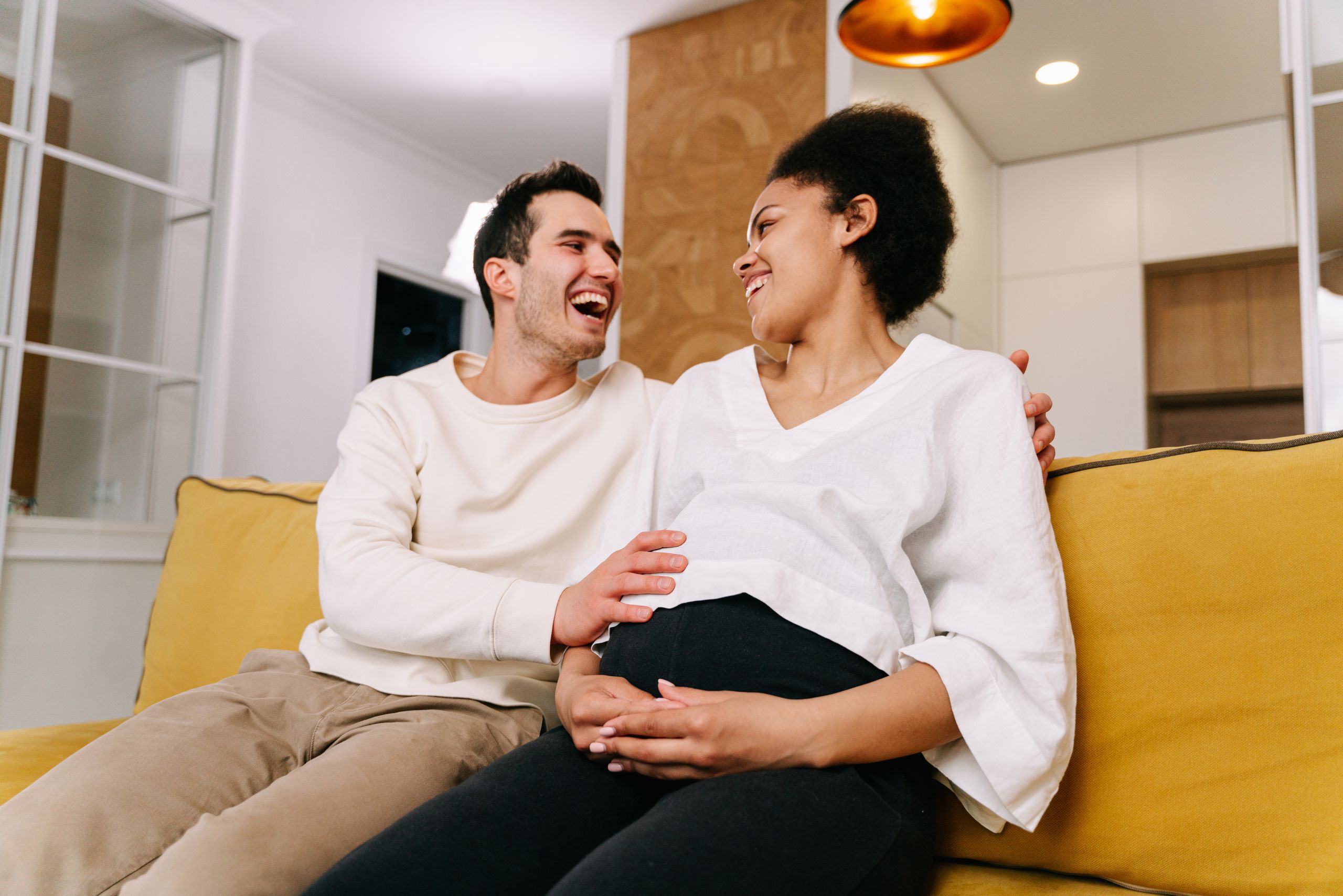 Cheerful couple enjoying a moment of happiness on a sofa during pregnancy. Indoor home setting.