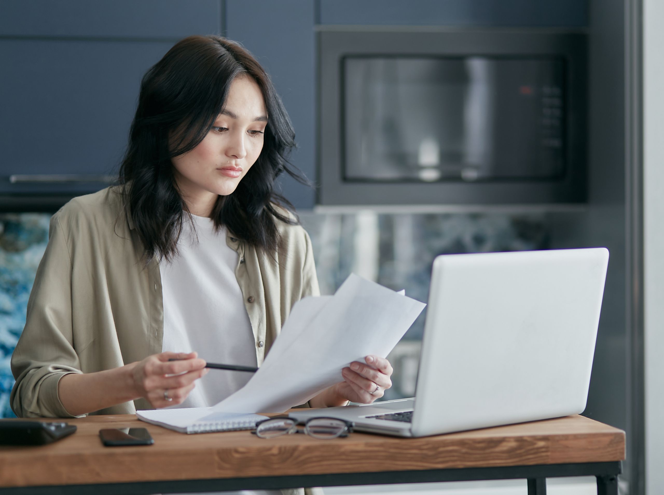 Concentrated woman reviewing documents and working on a laptop at home office setup in kitchen.