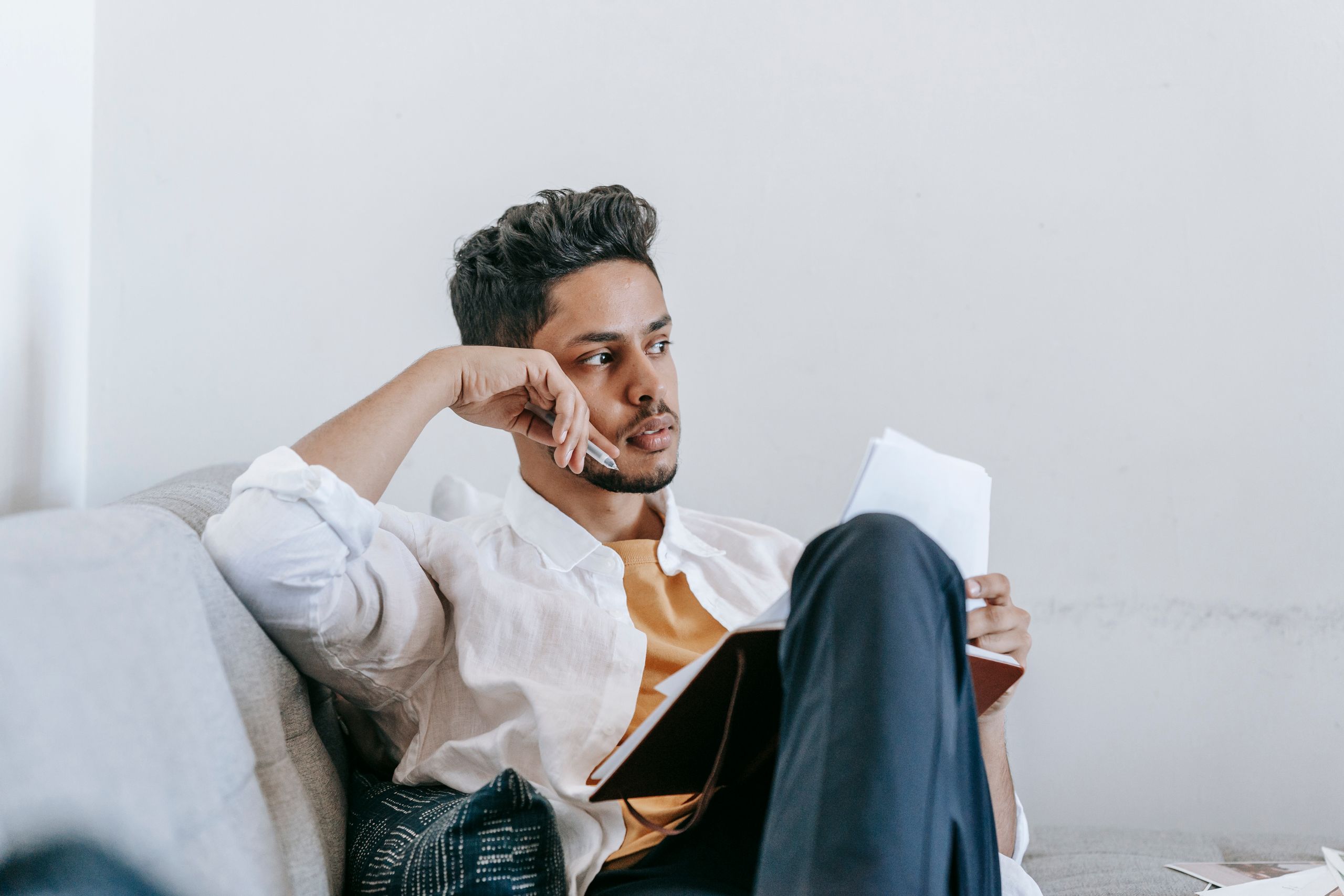Contemplative young ethnic male in casual wear reading notes in planner and looking away in thoughts while resting on comfortable couch