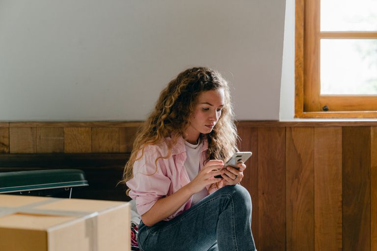 Content female in pink shirt and jeans sitting on bed with suitcase and carton box and browsing cellphone while moving in new apartment
