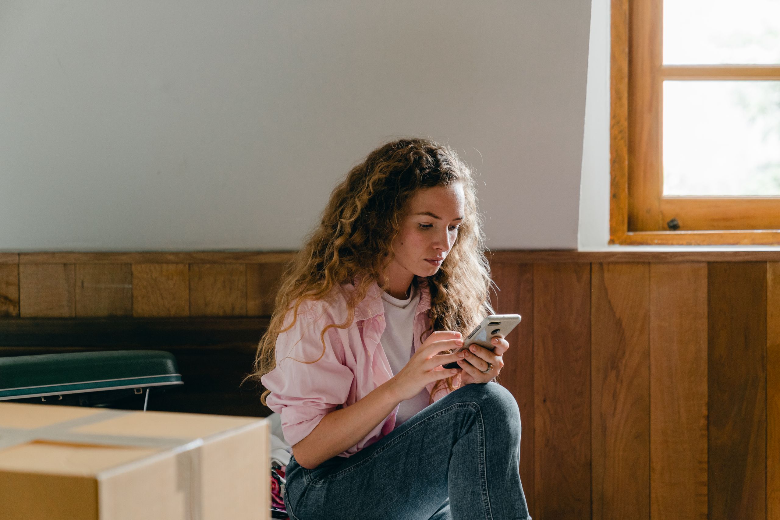 Content female in pink shirt and jeans sitting on bed with suitcase and carton box and browsing cellphone while moving in new apartment