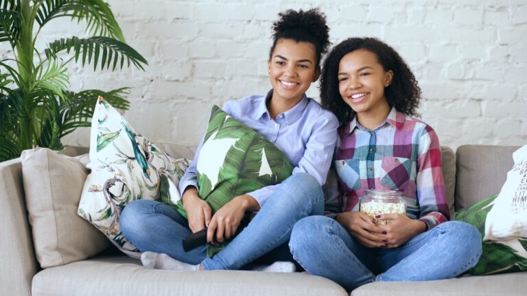 Two smiling young women watching television with popcorn.