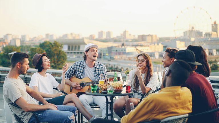 Group of friends having fun and playing guitar at a rooftop party with snacks and drinks.