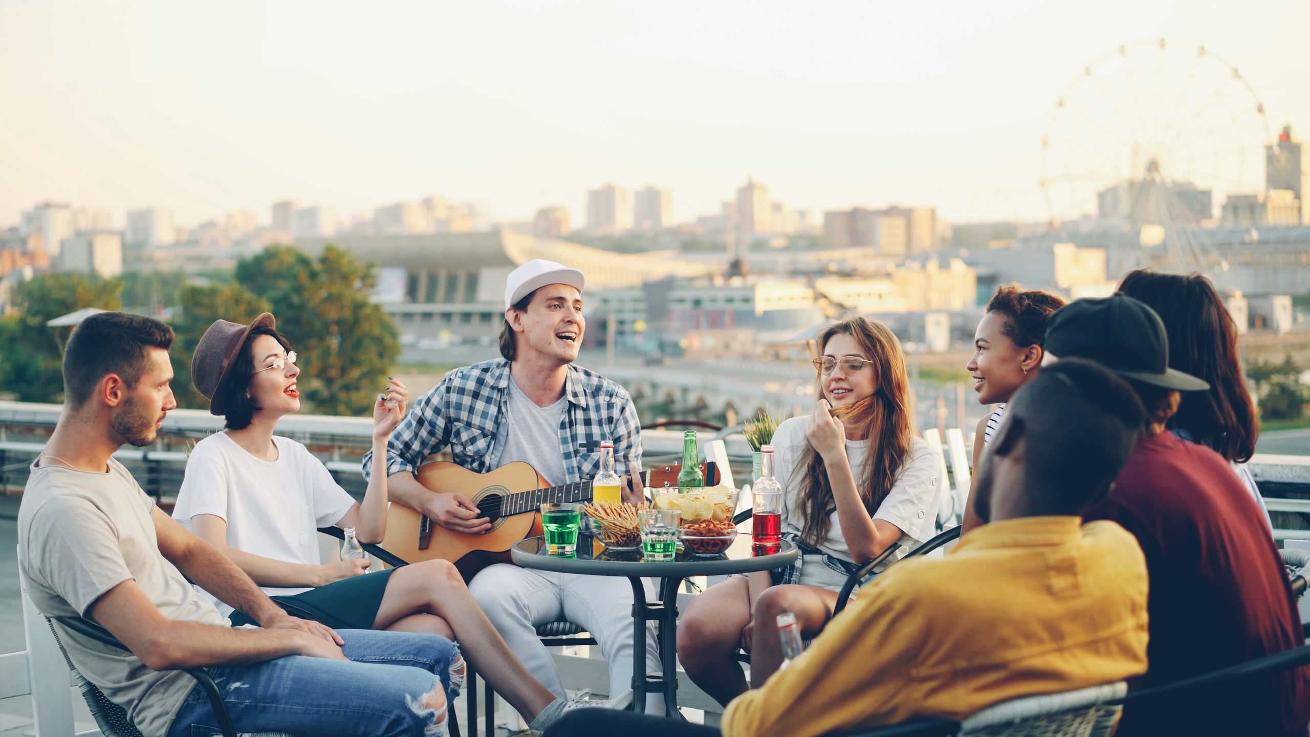 Group of friends having fun and playing guitar at a rooftop party with snacks and drinks.