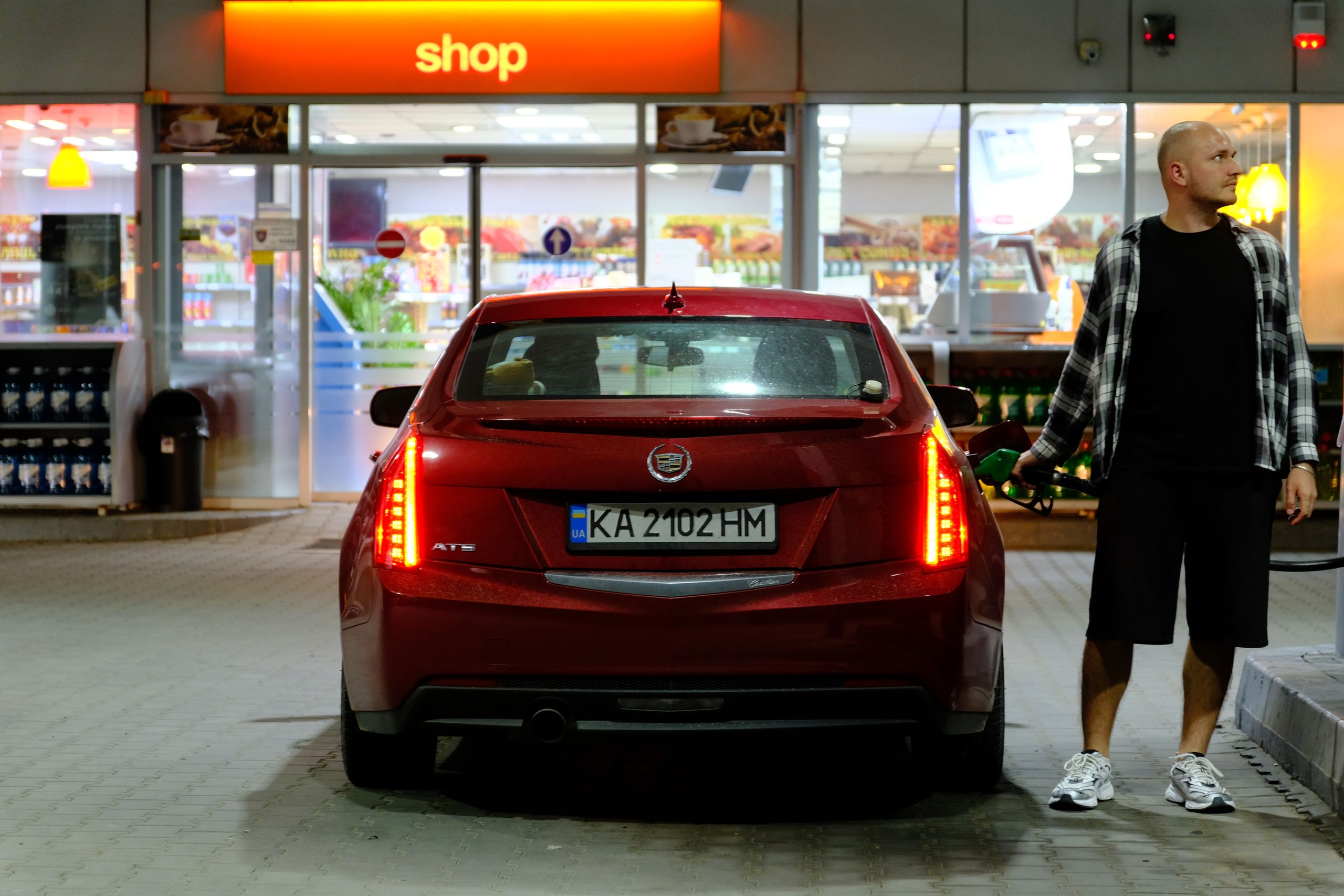 Man fueling a red car at a night-time gas station with a convenience shop in the background.