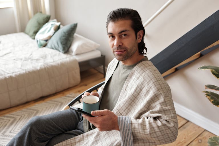Man in a comfortable home setting enjoying a cup of coffee while relaxing with a mobile device.