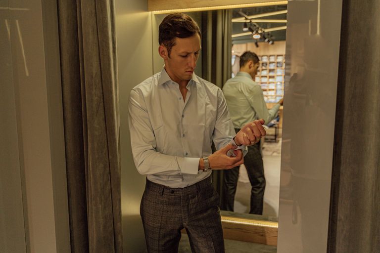 Man in stylish outfit adjusting shirt cuff in fitting room with mirror reflection.